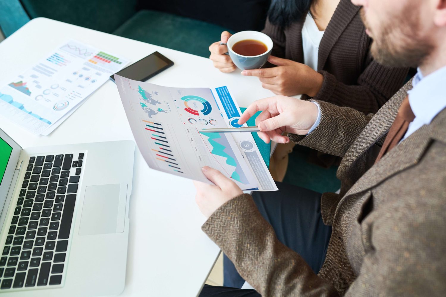 High angle crop shot of two contemporary business people holding graphs and charts while discussing strategy during meeting in cafe, laptop and documents on table